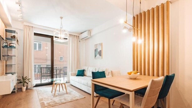 A modern living room with a wooden dining table, a sofa with teal cushions, and a large window with a view of the city. - Blue Lake Apartments (Ohrid)
