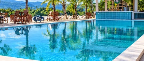 A clear swimming pool with lounge chairs and a bar area, surrounded by palm trees and a mountain view.