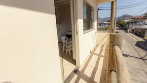 A narrow balcony with a wooden railing, a checkered table, and a chair, overlooking a street with buildings and mountains in the distance. - Apto c Varanda Garagem e Vista das Montanhas (Angra dos Reis)