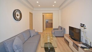 A modern living room with a glass coffee table, two grey sofas, a flat-screen TV, and a kitchen visible through an open doorway.