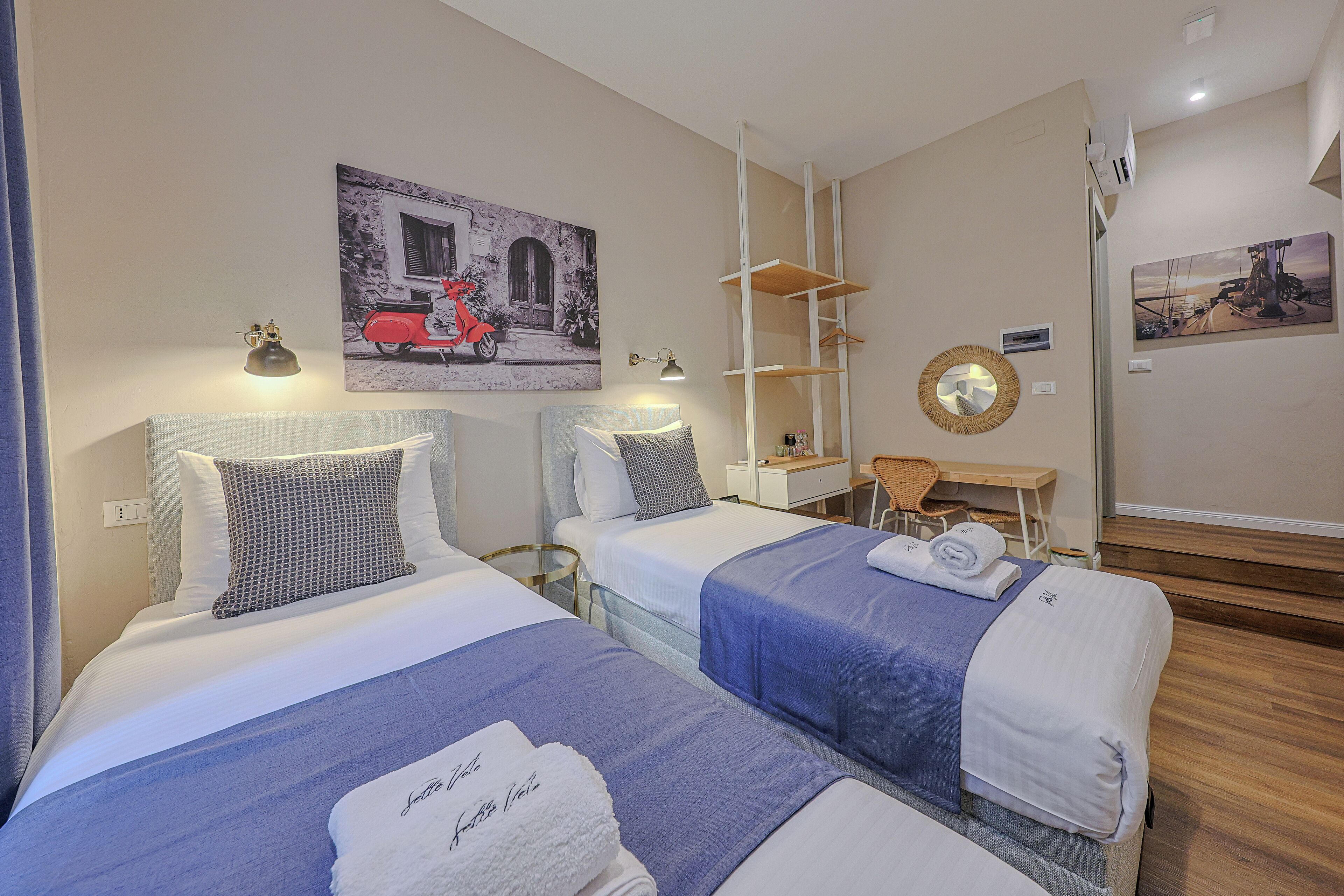A hotel room with two beds, a desk, and a chair. There are paintings on the walls and a mirror on the wall.