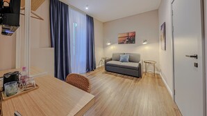 A modern hotel room with a wooden floor, a grey sofa, a dining table with a tray of items, a wicker chair, and a large window with curtains. - Sette Vele Rooms  (Trieste)