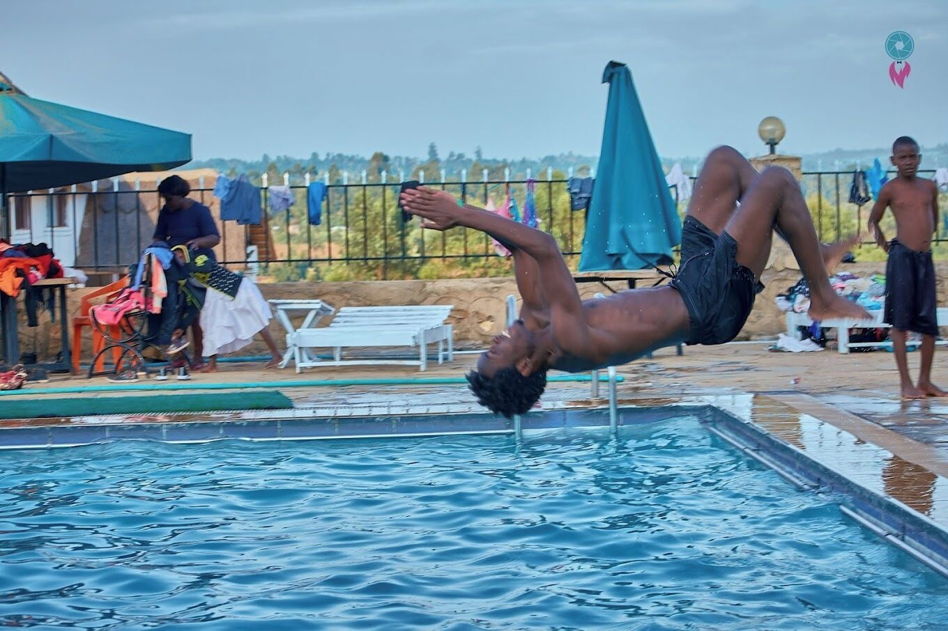 A man performing a backflip in a swimming pool with lounge chairs and umbrellas in the background.
