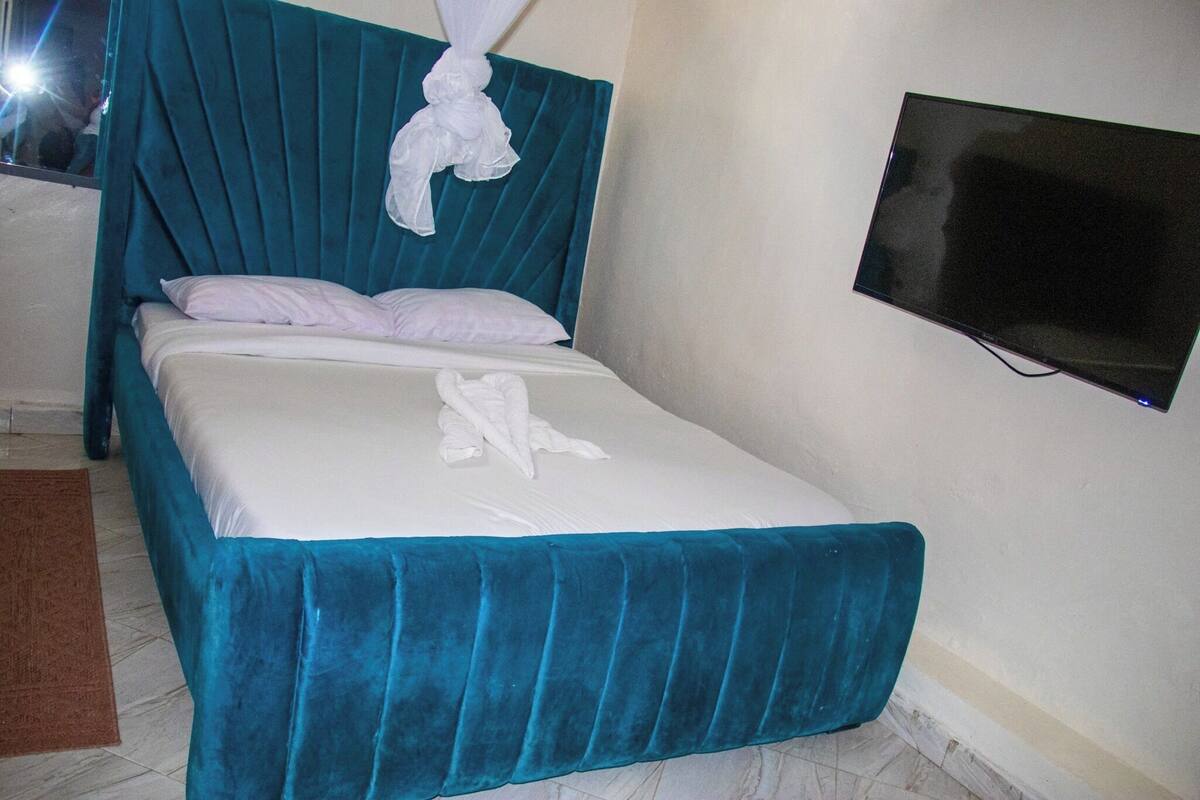 Una cama bien hecha con una colcha verde azulada y almohadas, una manta blanca y una toalla blanca sobre la cama. Un televisor de pantalla plana está montado en la pared a la derecha de la cama.