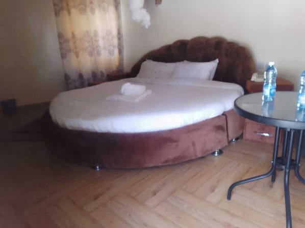 Ein Bett mit braunem Kopfteil, ein runder Tisch mit Wasserflaschen und ein Holzboden.