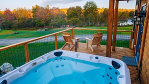 A hot tub on a wooden deck with chairs, overlooking a grassy area and trees.