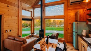A cozy cabin interior with a large window, wooden walls, a dining table, a sofa, and a kitchen area.