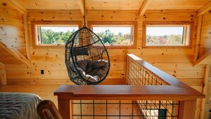A wooden cabin interior with a hanging chair, a wooden table, and two windows offering a view of trees.