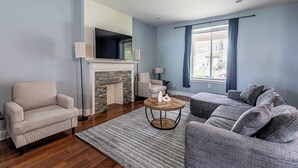 A living room with a fireplace, a sofa, a chair, and a TV mounted on the wall. - Spacious Modern 7BR Retreat ~ 3 Story Gem in Pittsburgh (Pittsburgh)