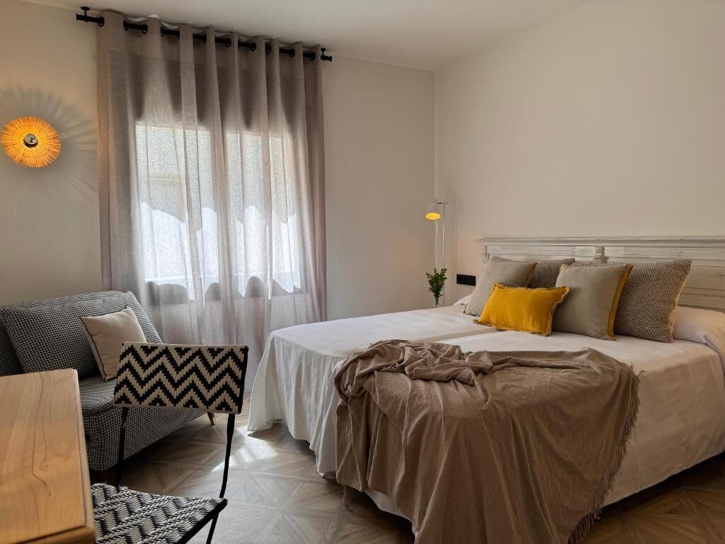 A bedroom with a bed, a sofa, a chair, a lamp, and a wall-mounted light fixture.