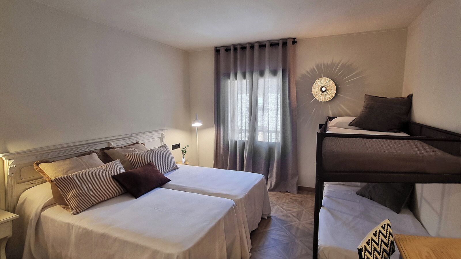 A hotel room with two single beds, a bunk bed, a bedside lamp, a decorative wall piece, and a window with curtains.