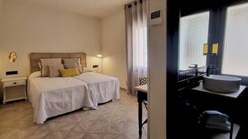A hotel room with a double bed, bedside tables, a small table with a chair, a window with curtains, and a bathroom with a sink and mirror.