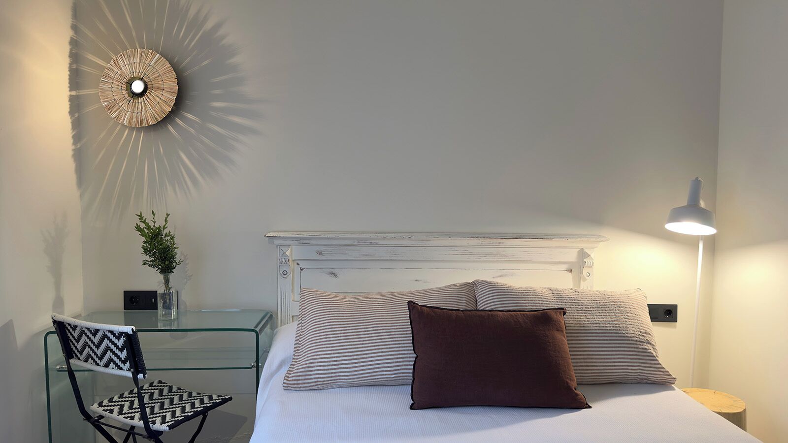 A bed with striped bedding and a brown pillow, a glass table with a chair, a wall-mounted lamp, and a decorative wall piece.
