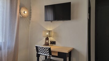 Una habitación con una televisión de pantalla plana montada en la pared, un pequeño escritorio de madera, una silla con respaldo estampado en blanco y negro, y una lámpara fijada a la pared.
