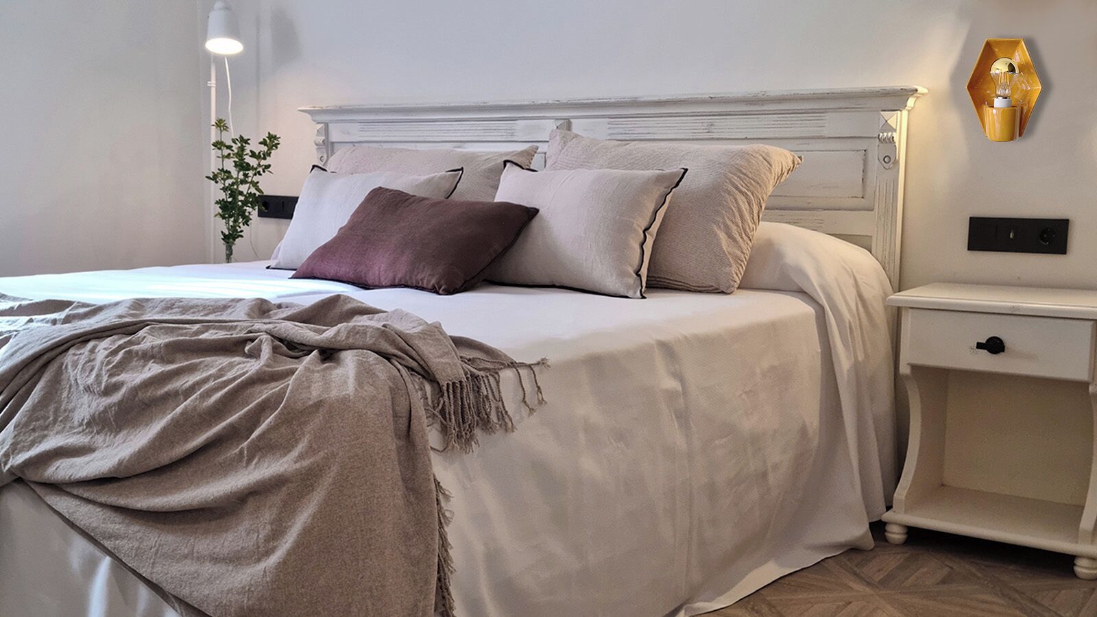 A neatly made bed with a white headboard, a bedside table with a lamp, and a wall-mounted light fixture.