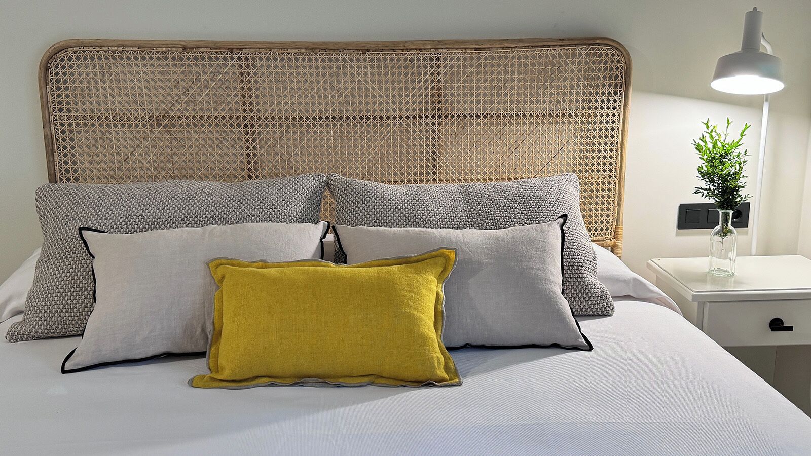 Una cama con cabecera de mimbre, varios almohadones, una mesita de noche con lámpara y una planta pequeña.