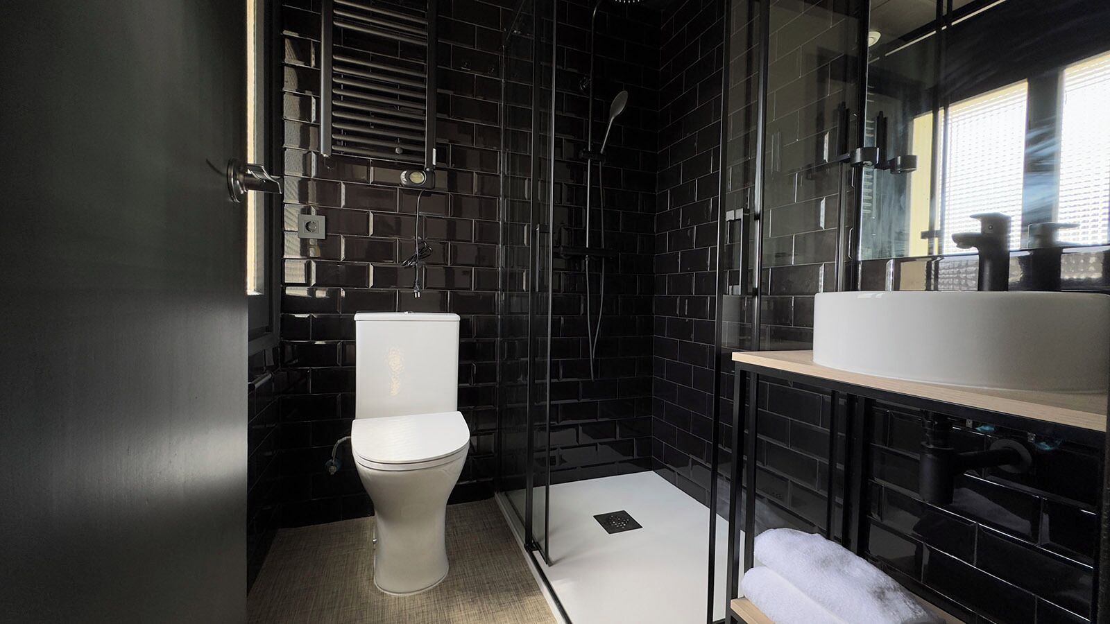 Un bany modern amb dutxa revestida de rajoles negres, vàter blanc i un lavabo amb mirall.