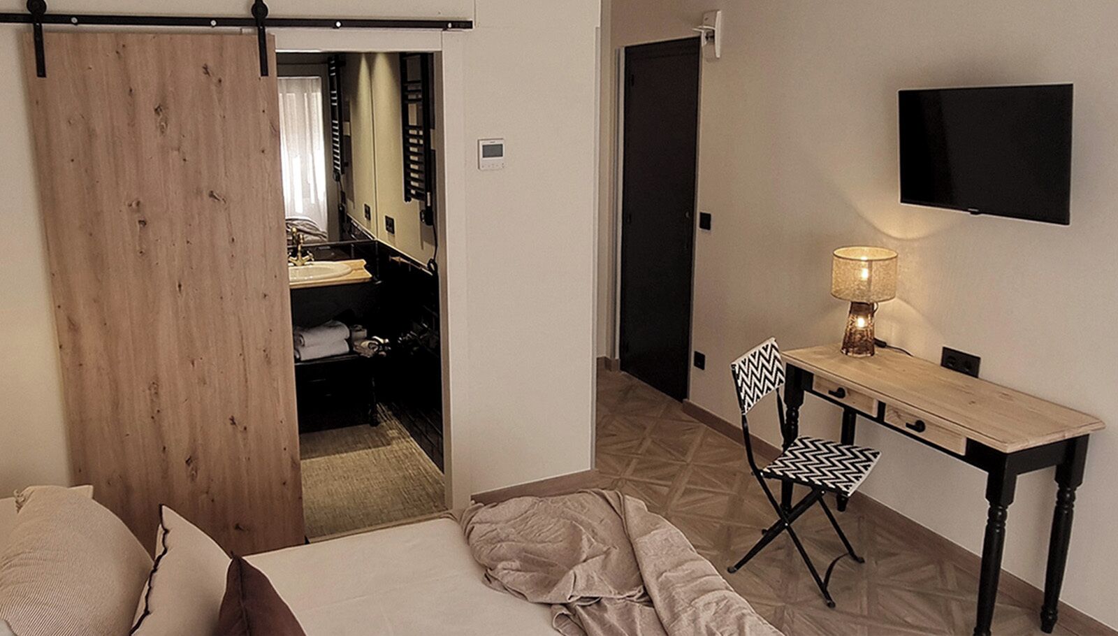 Una habitación de hotel con cama, un escritorio con silla, un televisor montado en la pared y un armario.