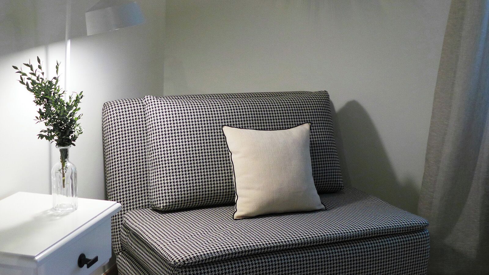 A houndstooth-patterned sofa with a single pillow, a white bedside table with a vase of green plants, and a lamp.