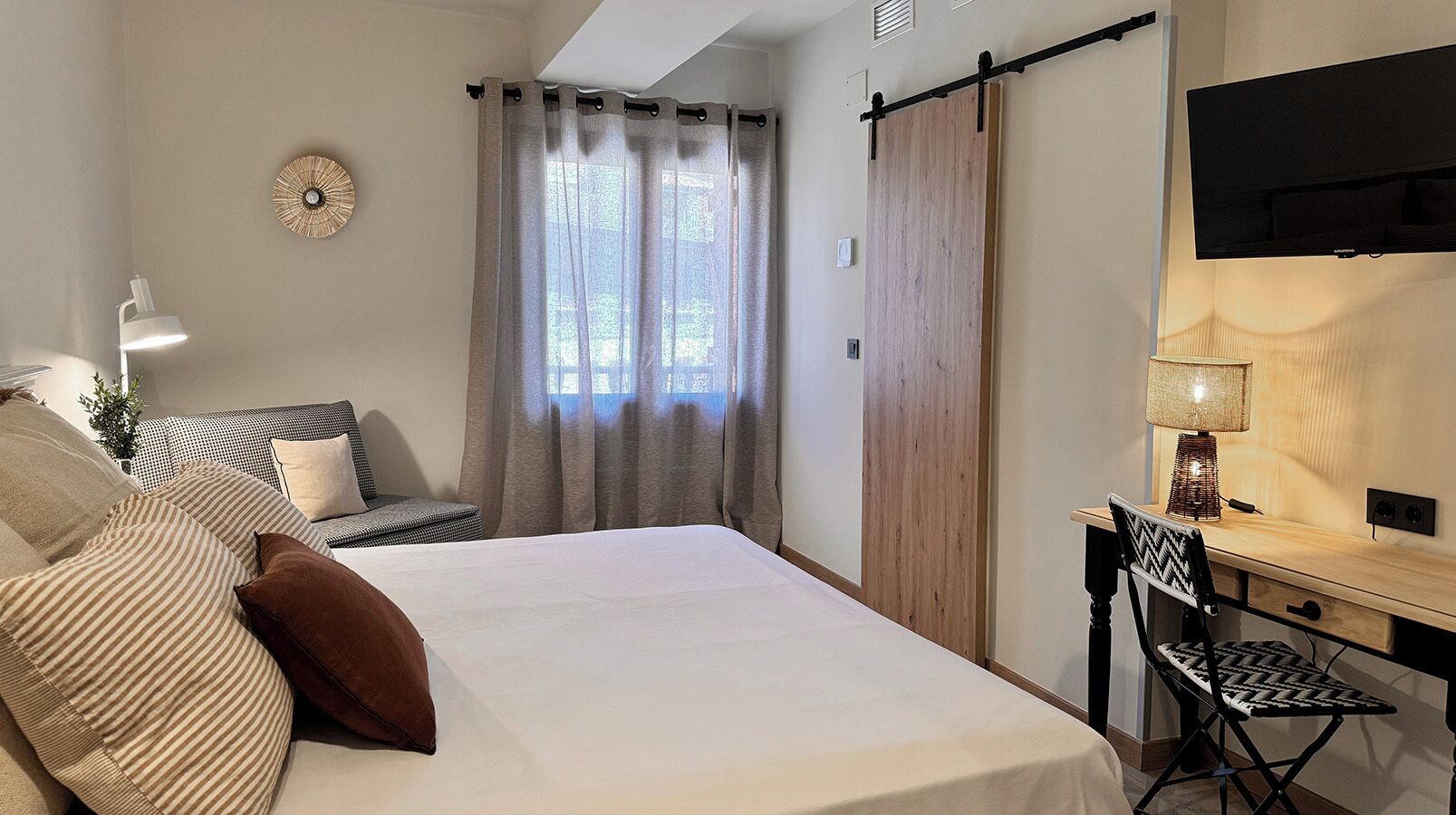 Un dormitorio con una cama, un escritorio con silla, un televisor, una ventana con cortinas y un reloj de pared.