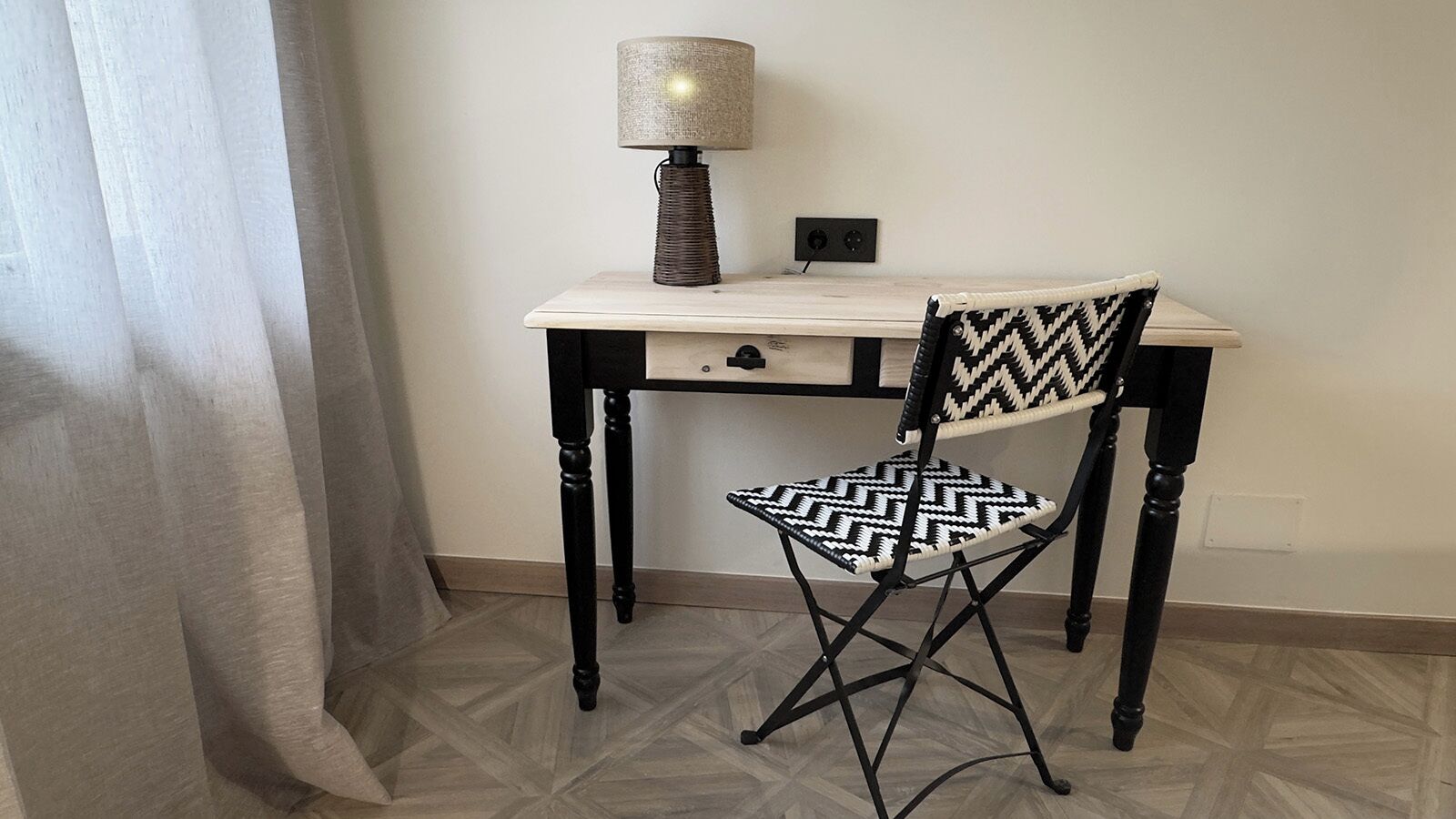 Una habitación con un escritorio de madera, una silla con estampado blanco y negro, y una lámpara.