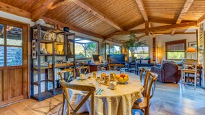 A spacious room with a wooden ceiling, a dining table with chairs, a bookshelf, and a large window.