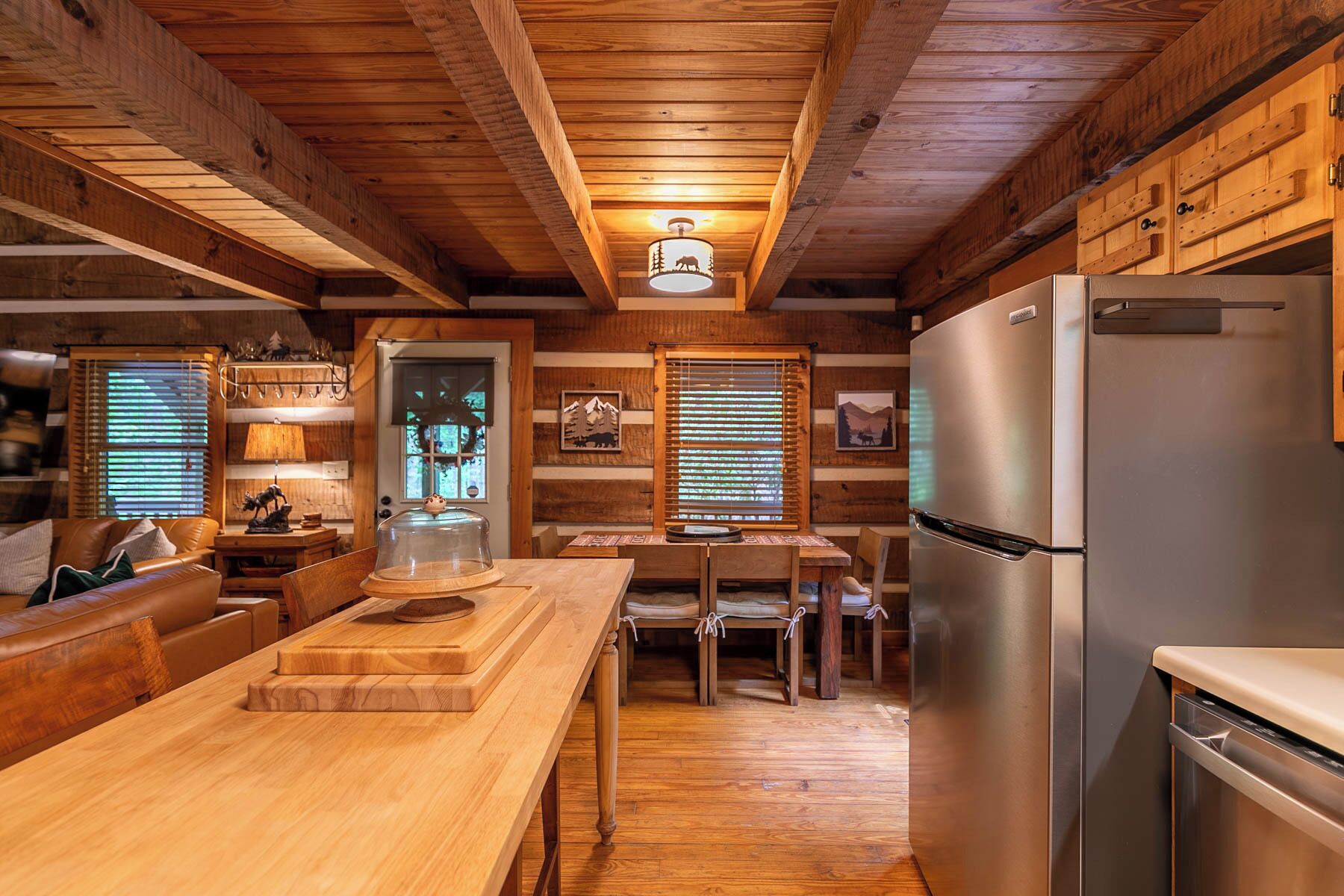 A cozy cabin interior with wooden walls, a dining area, a kitchen with a stainless steel refrigerator, and a wooden ceiling.