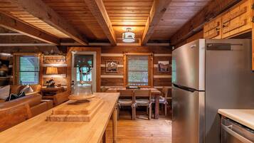 A cozy cabin interior with wooden walls, a dining area, a kitchen with a stainless steel refrigerator, and a wooden ceiling.