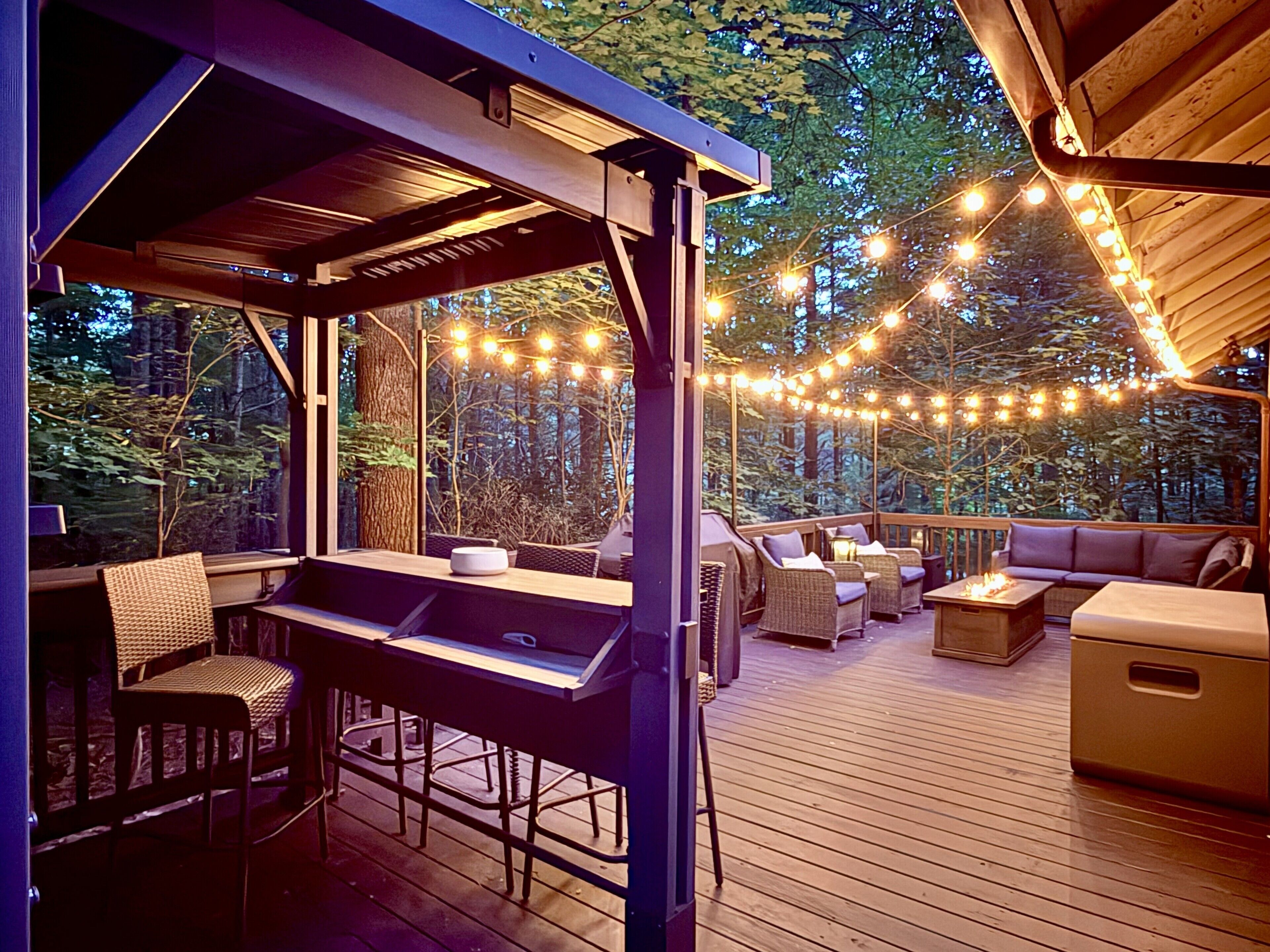 An outdoor deck with a wooden table, chairs, and a sofa, surrounded by string lights and a view of trees.