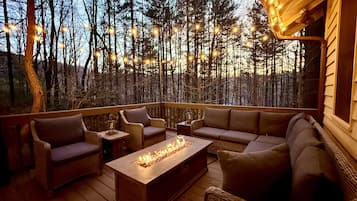 A wooden deck with wicker furniture, a fire feature, and string lights against a forest backdrop.