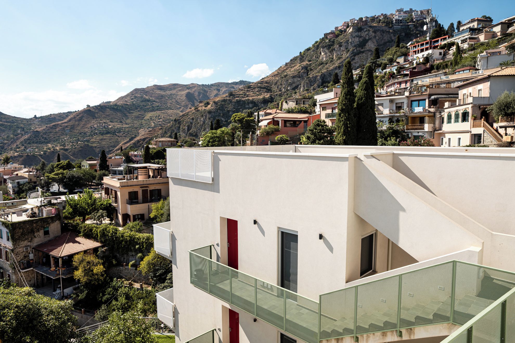 Un edificio moderno con un balcone in vetro, immerso in un paesaggio collinare e circondato da altri edifici residenziali.