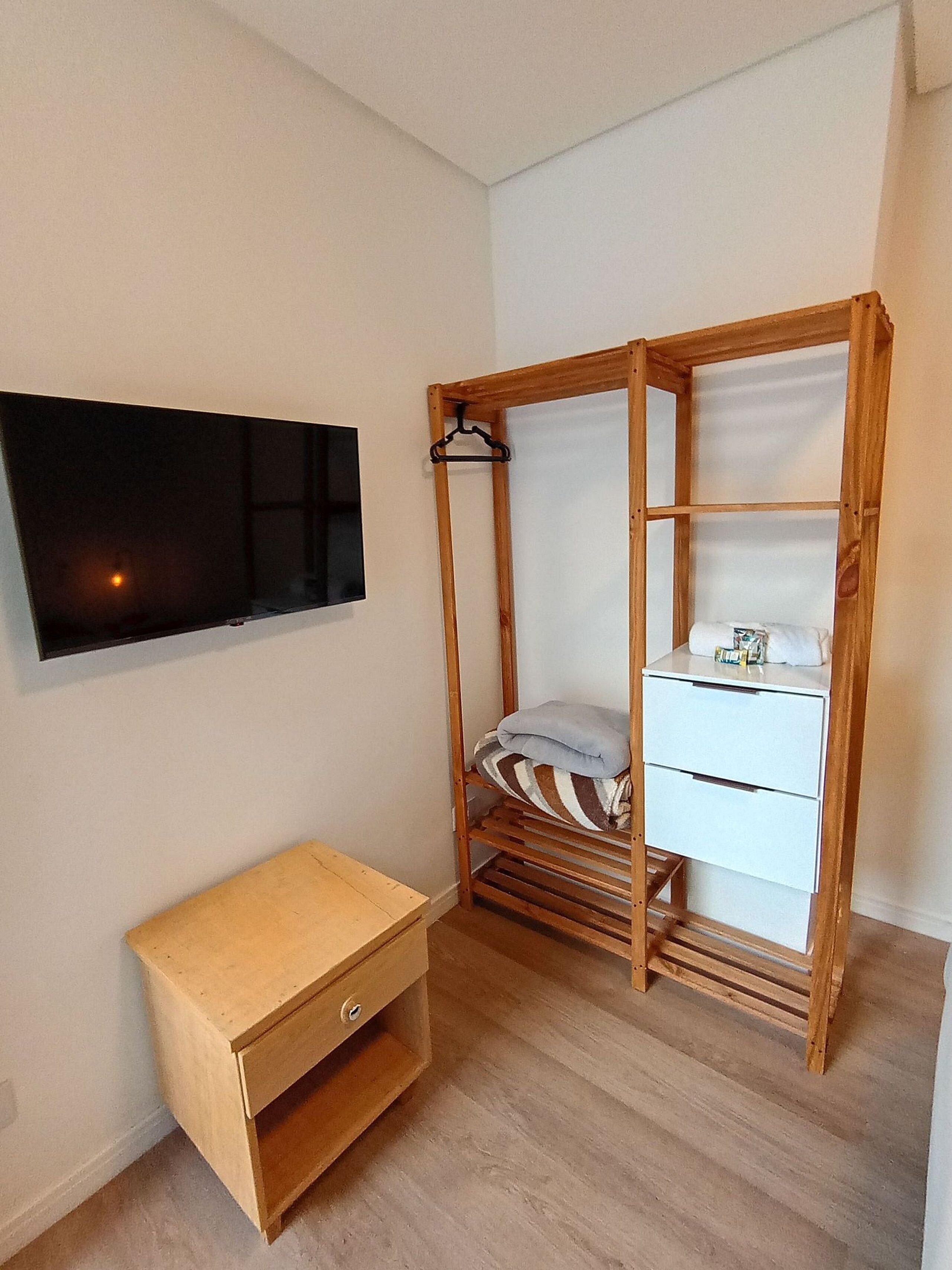 Un dormitorio con un armario de madera, una mesita de noche pequeña y un televisor instalado en la pared.