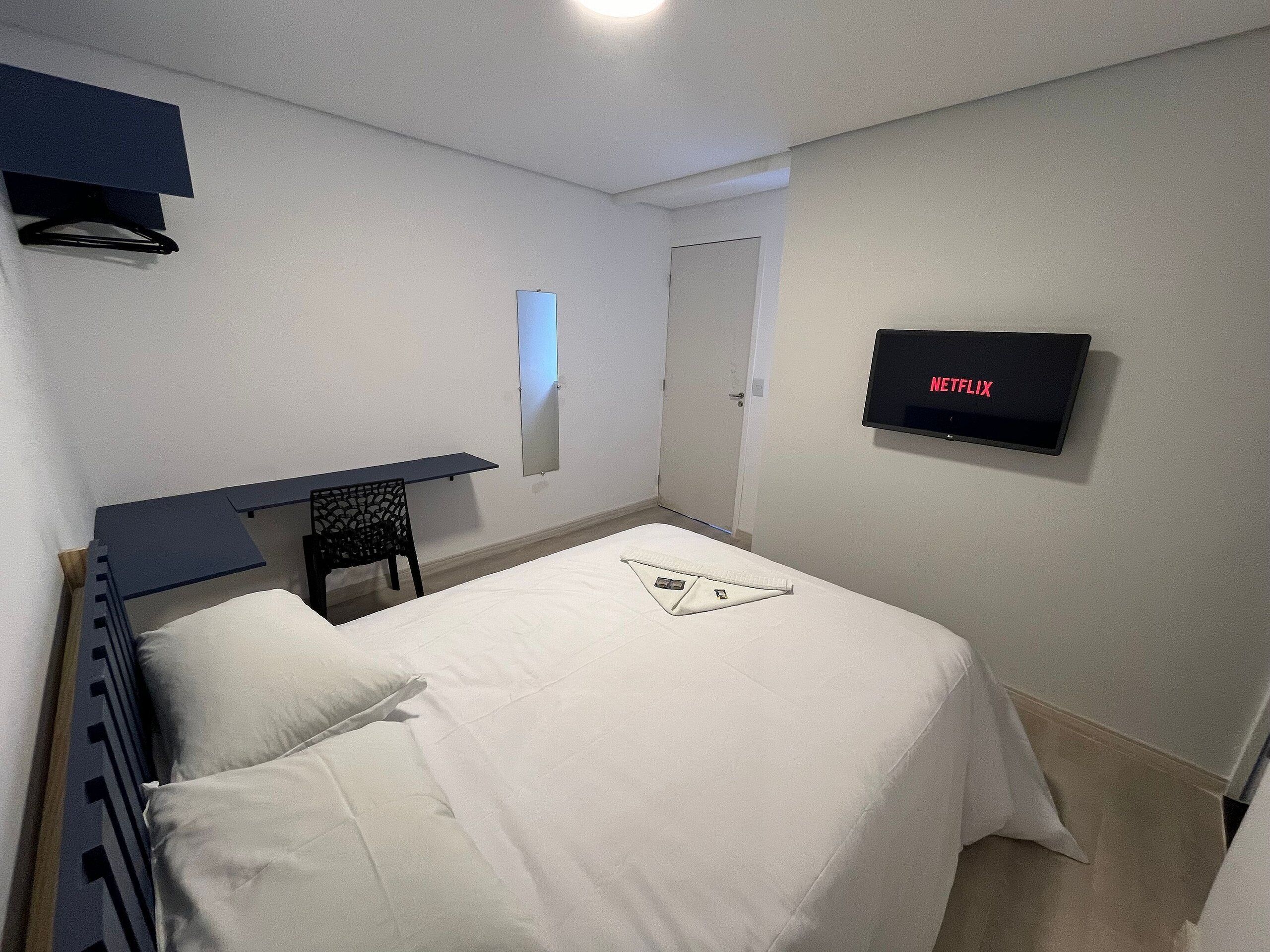 A bedroom with a bed, a desk, a chair, a TV mounted on the wall, and a door.