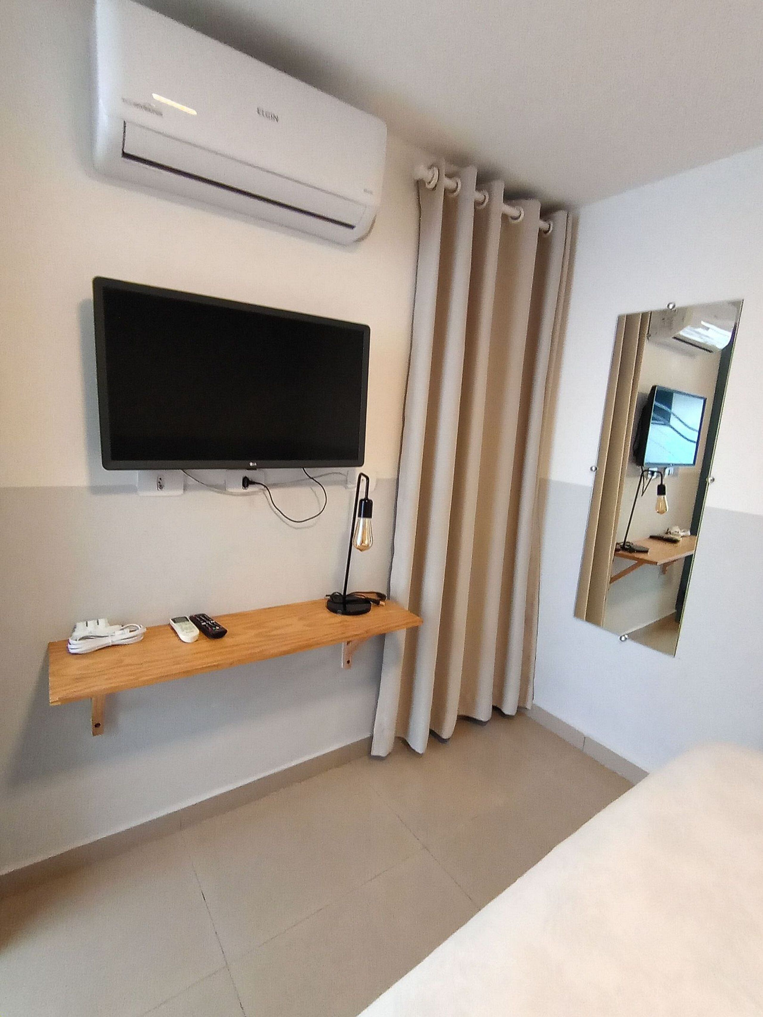 A hotel room with a flat-screen TV mounted on the wall, a wooden shelf with a lamp, and a mirror reflecting the room.