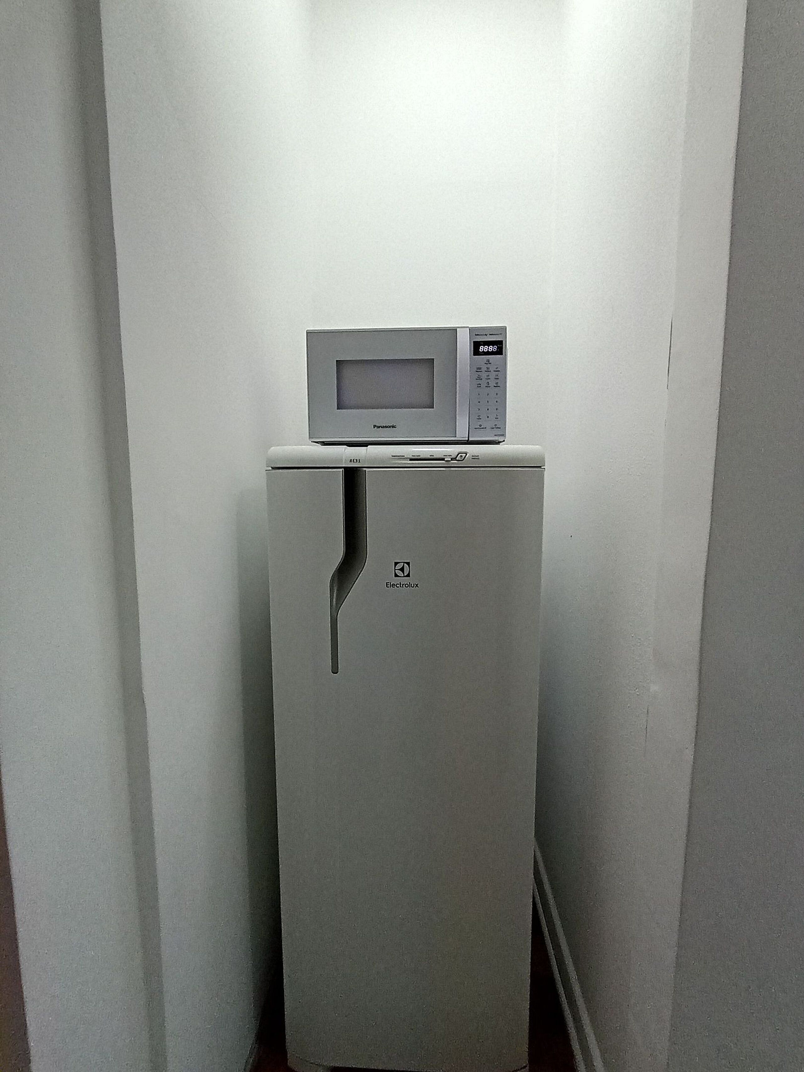 Een compacte koelkast met een ingebouwde magnetron erboven, geplaatst in een smalle gang met eenvoudige wanden.