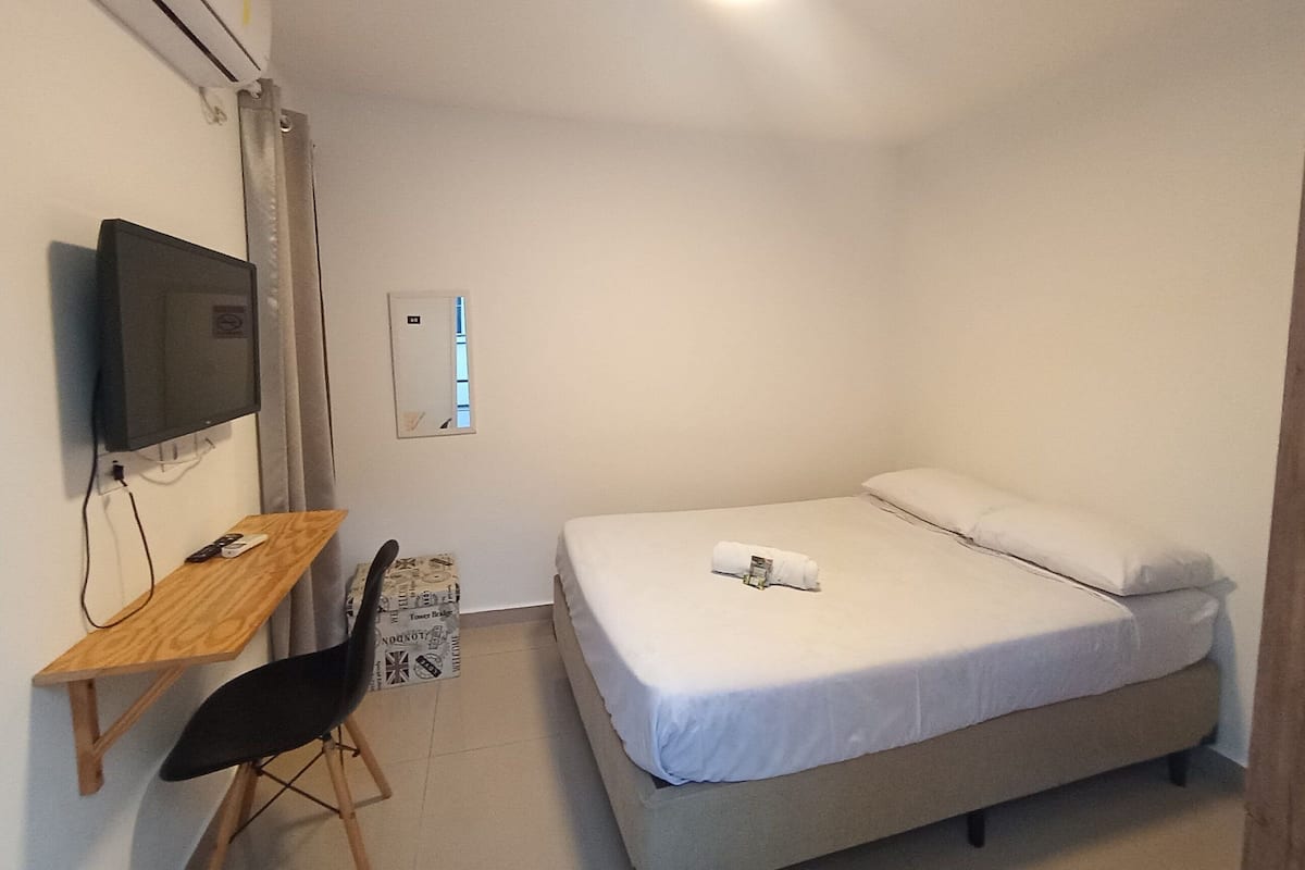 Ein kleiner, sauberer Raum mit einem Bett, einem Schreibtisch mit Stuhl, einem Fernseher und einem Spiegel.