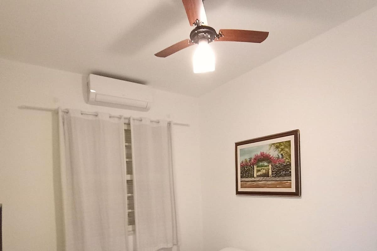 Ein Schlafzimmer mit Bett, weißen Vorhängen, Deckenventilator, Klimaanlage und einem gerahmten Bild an der Wand.