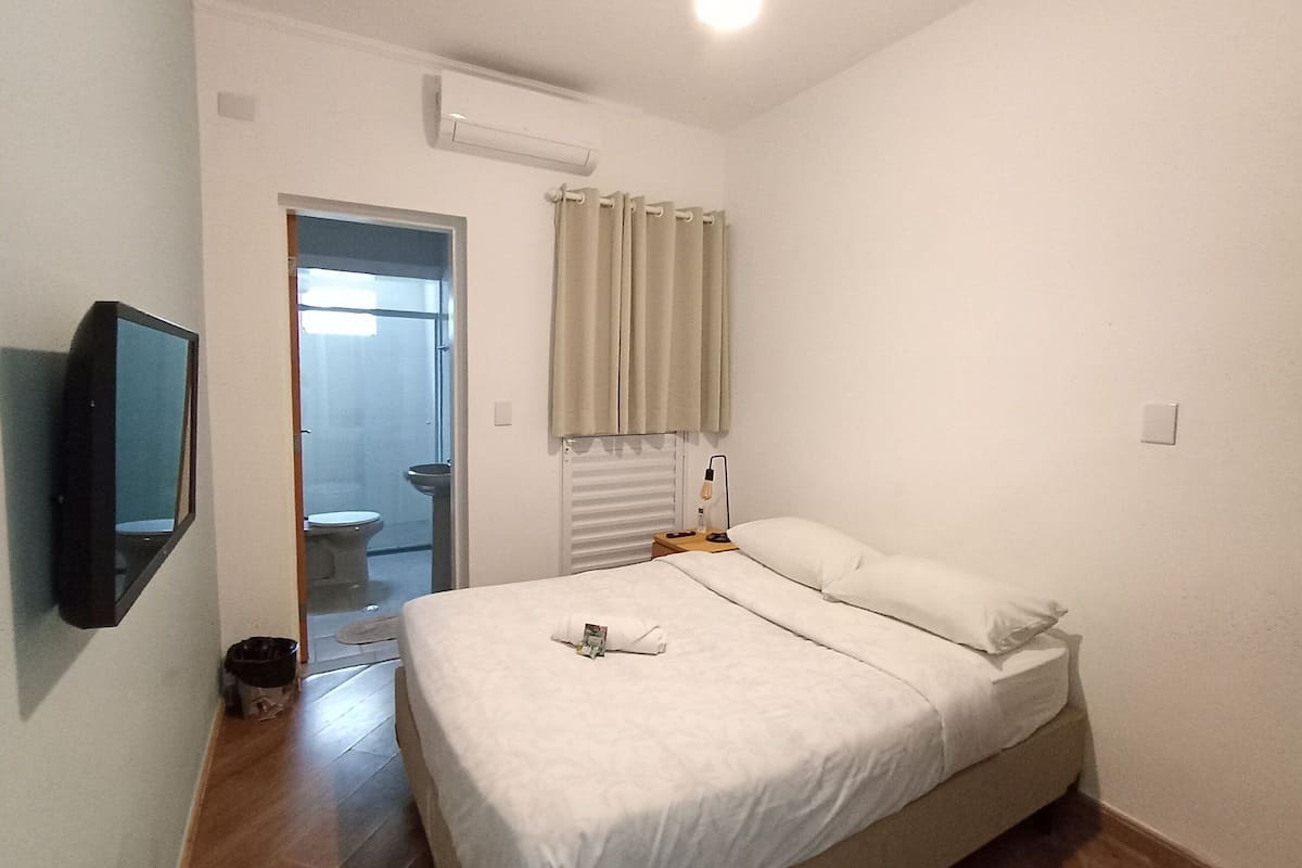Ein Schlafzimmer mit einem Bett, einem an der Wand montierten Fernseher, einem Fenster mit Vorhängen und einer Klimaanlage.