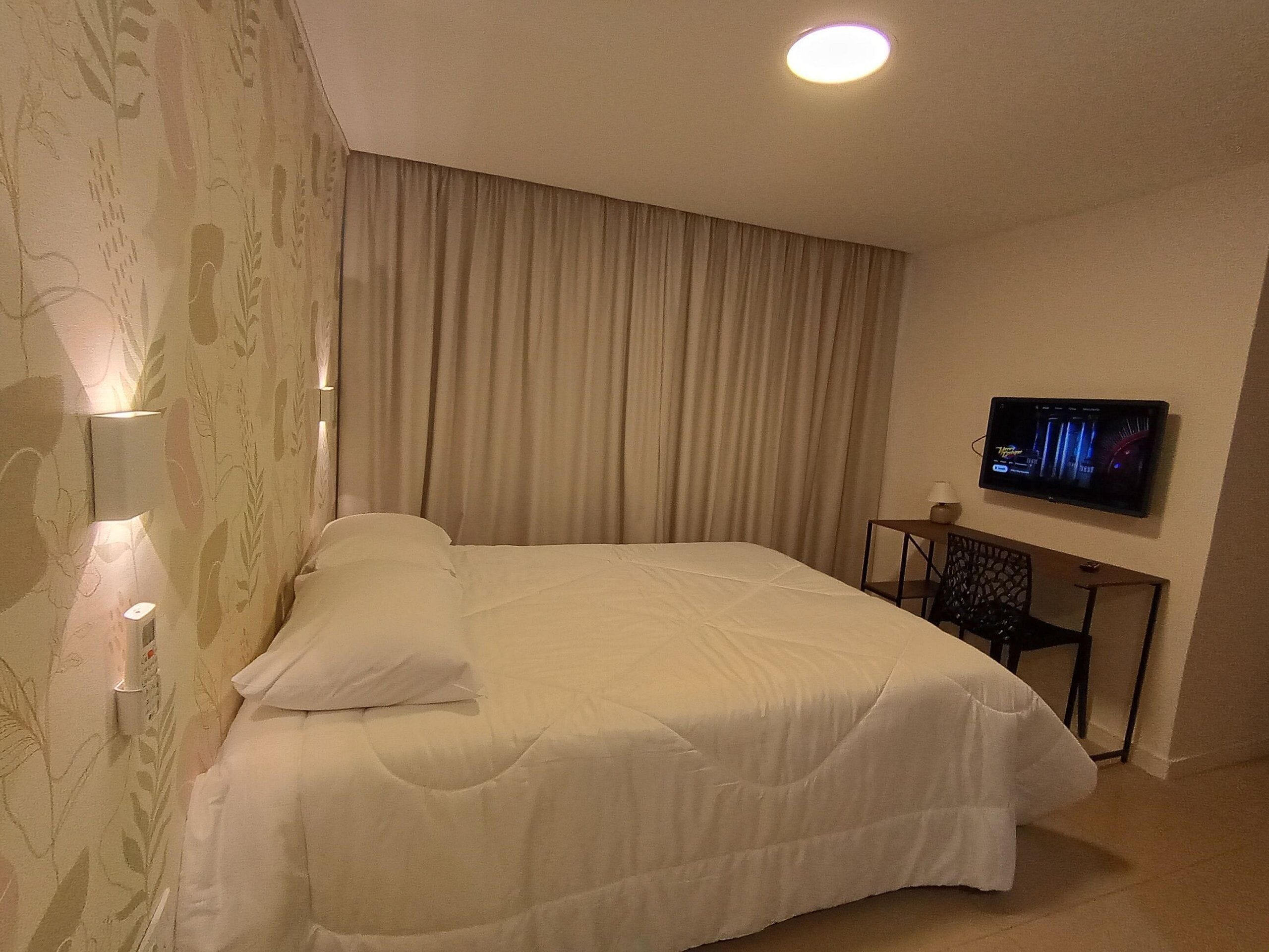 Hotellihuone, jossa on suuri sänky, seinälle asennettu televisio ja ikkuna, jossa on verhot.