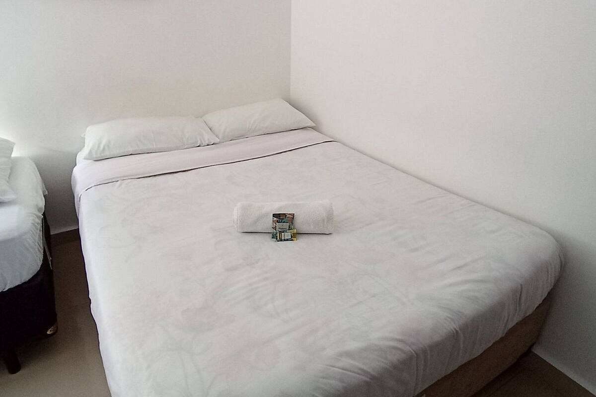 Ein ordentlich bezogenes Bett mit weißen Laken, ein kleiner Nachttisch mit Lampe und ein Blumenbild an der Wand.