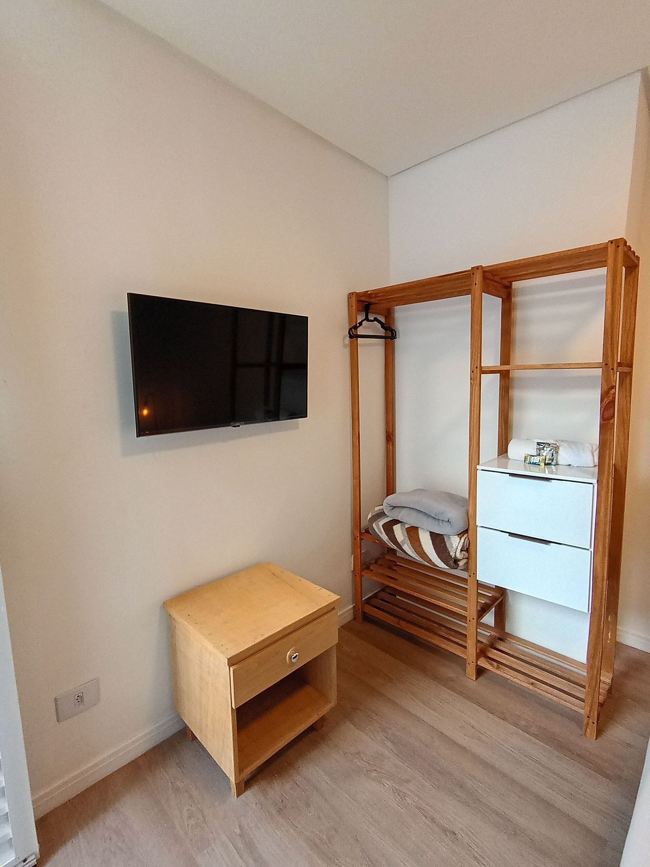A small, neatly organized room with a wooden bed, a bedside table, a TV mounted on the wall, and a window.