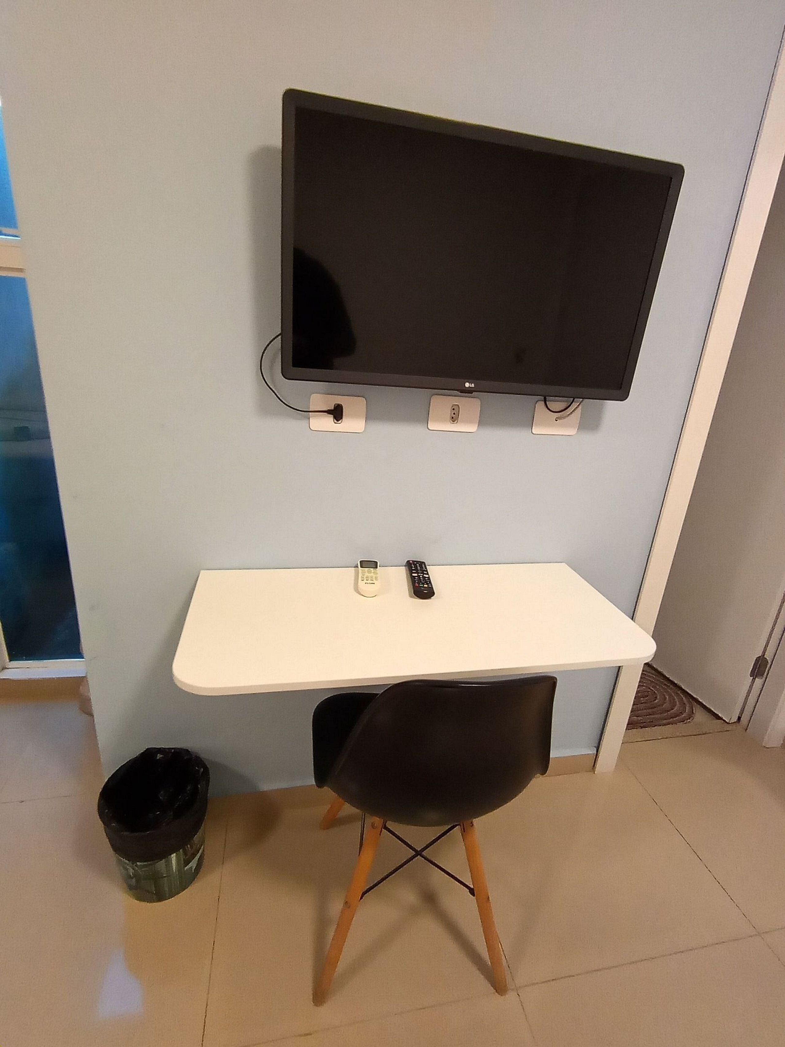 A small room with a white desk, a black chair, a television mounted on the wall, and a trash bin.