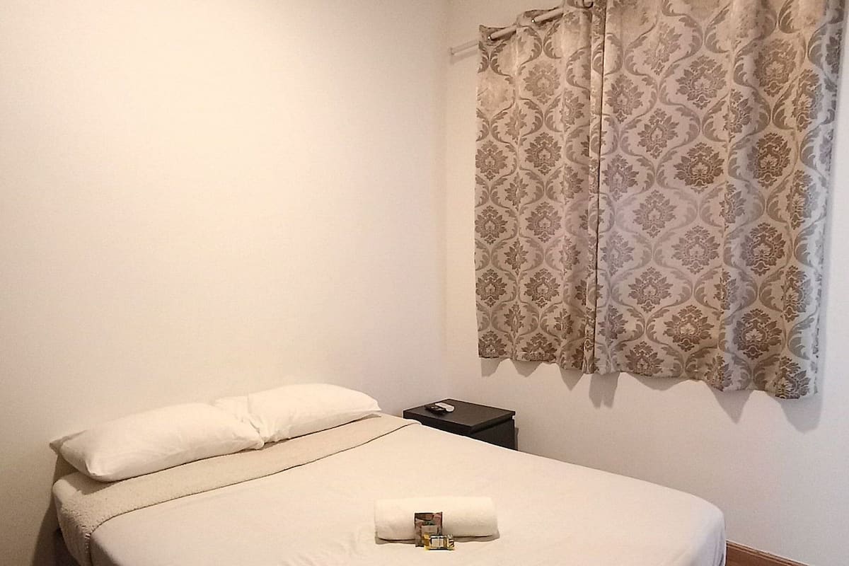 Ein ordentlich bezogenes Bett mit weißer Bettdecke und einem aufgerollten Handtuch in der Mitte, ein kleiner Nachttisch mit Fernbedienung und ein fenster mit gemustertem Vorhang.