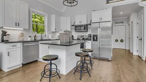 Una cocina moderna con gabinetes blancos, una isla central con taburetes, electrodomésticos de acero inoxidable y una araña.