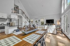 Une cuisine moderne avec des meubles blancs, un coin repas doté d’une table en bois et un escalier menant à une mezzanine.