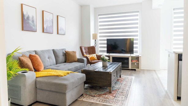 Una sala de estar moderna con un sofá seccional gris, una mesa de centro de madera y un televisor. Hay tres cuadros enmarcados en la pared, una planta en maceta y un ventanal con persianas.
