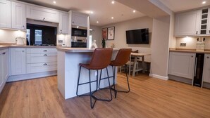 A modern kitchen with white cabinets, a central island with bar stools, a dining area with a TV, and wooden flooring. - Church Road Retreats - The Reef (Kessingland)