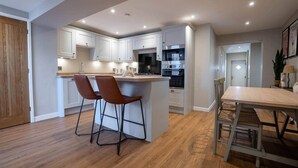 A modern kitchen with white cabinets, a central island with stools, and a dining area with a wooden table. - Church Road Retreats - The Reef (Kessingland)