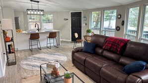 A spacious living room with a brown leather couch, a glass coffee table, and a dining area with a kitchen in the background.