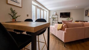 A modern living room with a wooden dining table, black chairs, a pink sofa, and a TV on the wall. - Church Road Retreats - Coral Cottage (Kessingland)